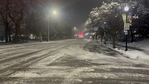A dark, icy street in the early morning. Streetlights reflect off the slick surface, with faint tire tracks visible. Hazardous conditions.